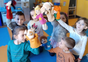 Grupa dzieci klęczy na podłodze i w wyciągniętych ku górze rękach trzymają lalki-pacynki.